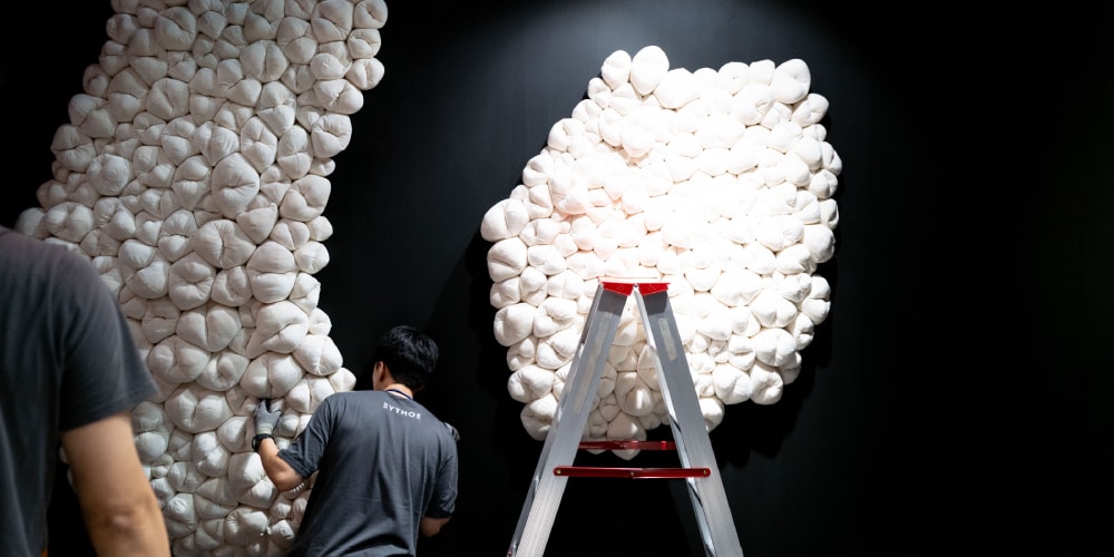 Installation d'art en coussins blancs sur mur noir, homme et échelle visibles.