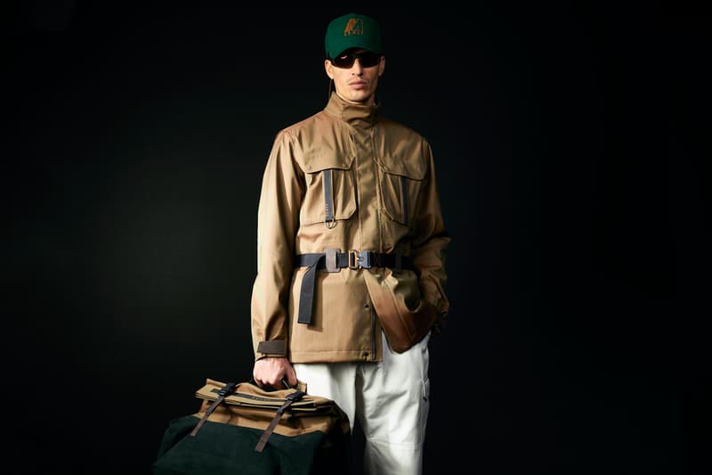 Homme en veste beige, casquette verte et lunettes de soleil, sac en main.